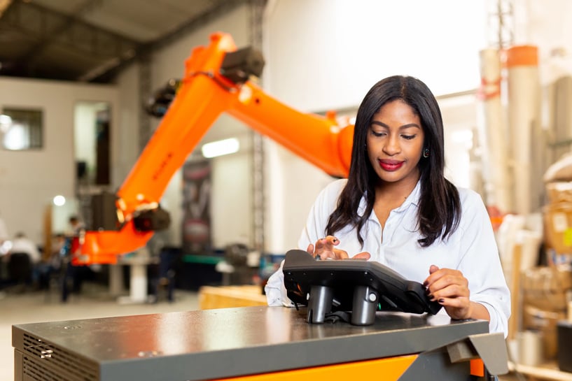 Una mujer en una fábrica utiliza una pantalla táctil para operar un sistema industrial, con un brazo robótico naranja en el fondo que simboliza la tecnología y la innovación en la manufactura