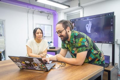  Two people collaborating at a table with a laptop decorated with stickers, while a screen in the background displays a graphic model