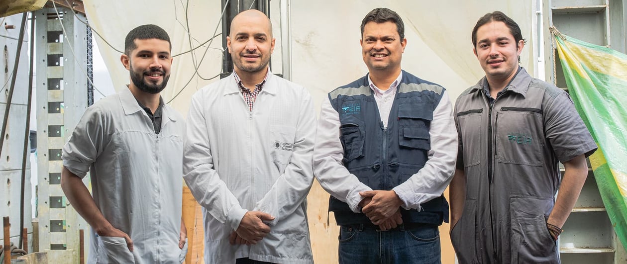 Cuatro hombres sonriendo y posando juntos, algunos con batas de laboratorio y otros con chalecos de trabajo.