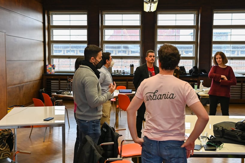 Grupo de personas conversando de pie en una sala con mesas y sillas, participando en una actividad colaborativa