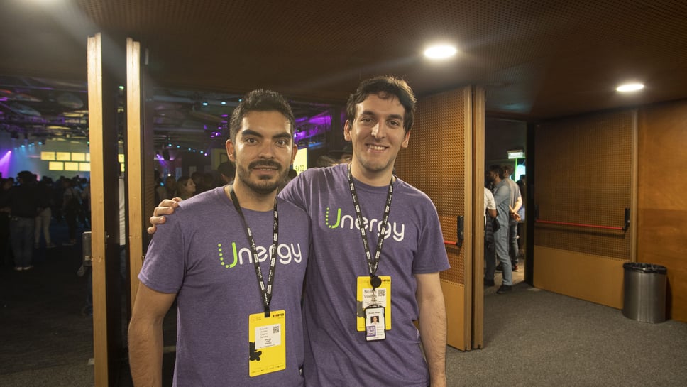 Dos personas en primer plano en un evento concurrido con iluminación colorida y un ambiente vibrante.