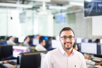 Hombre con gafas sonriendo en una oficina moderna, rodeado de estaciones de trabajo con monitores y compañeros en el fondo