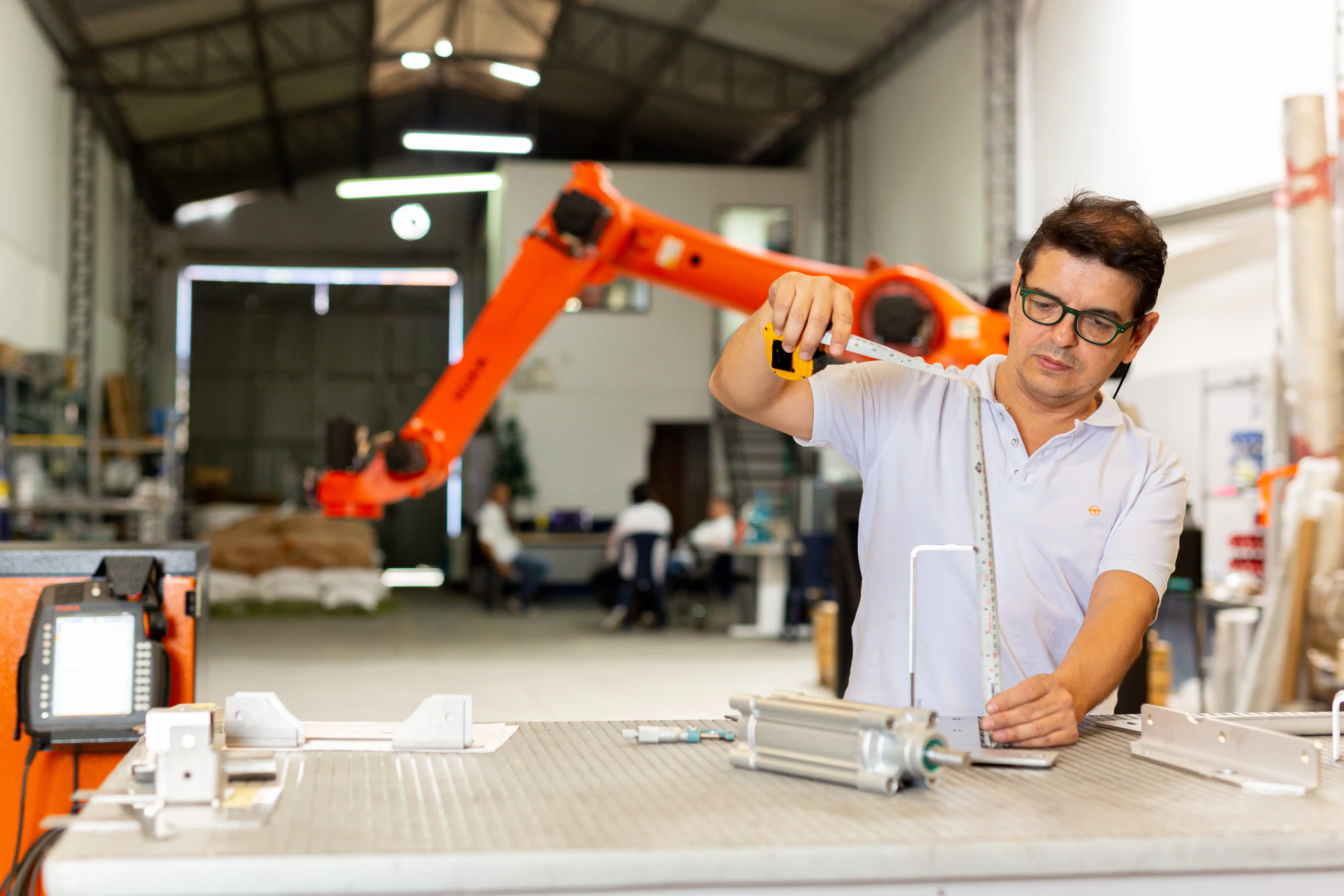 Un hombre en una fábrica mide piezas mecánicas con una cinta métrica en una mesa de trabajo, con un brazo robótico naranja visible en el fondo, representando un ambiente de manufactura y tecnología industrial.