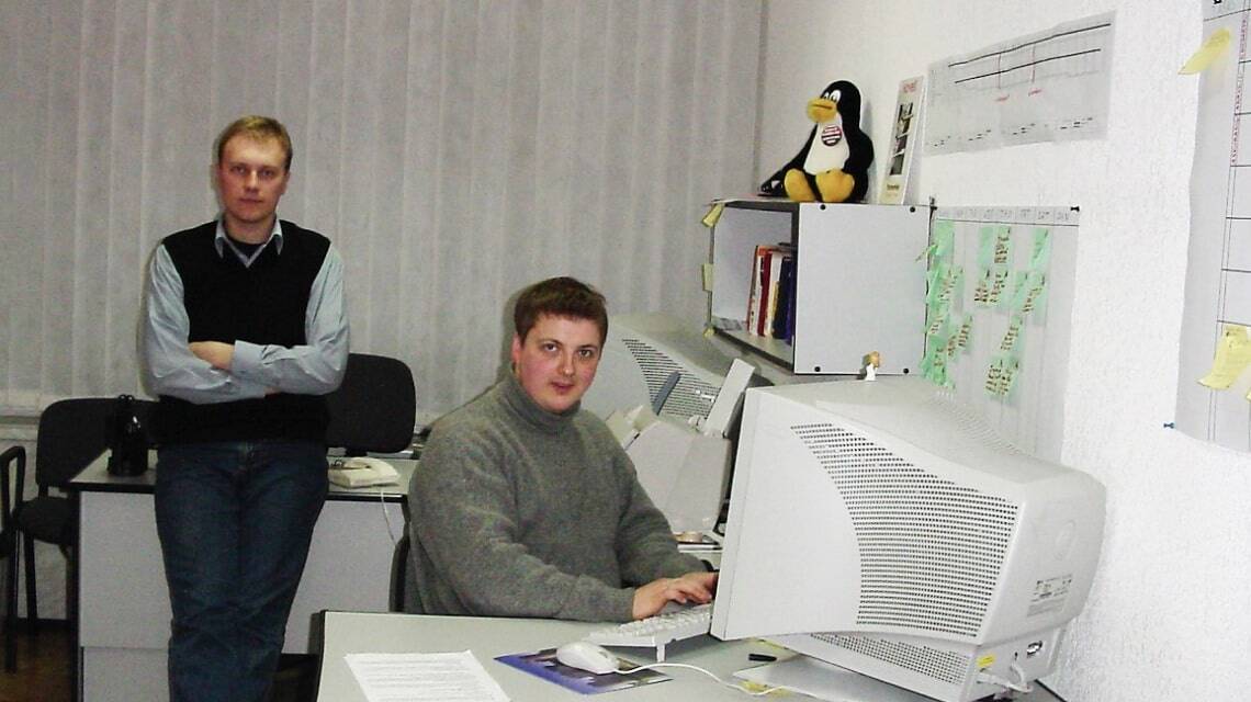 Dos hombres en una oficina con computadoras de escritorio antiguas, uno sentado trabajando y otro de pie junto a una estantería con un peluche de Linux.