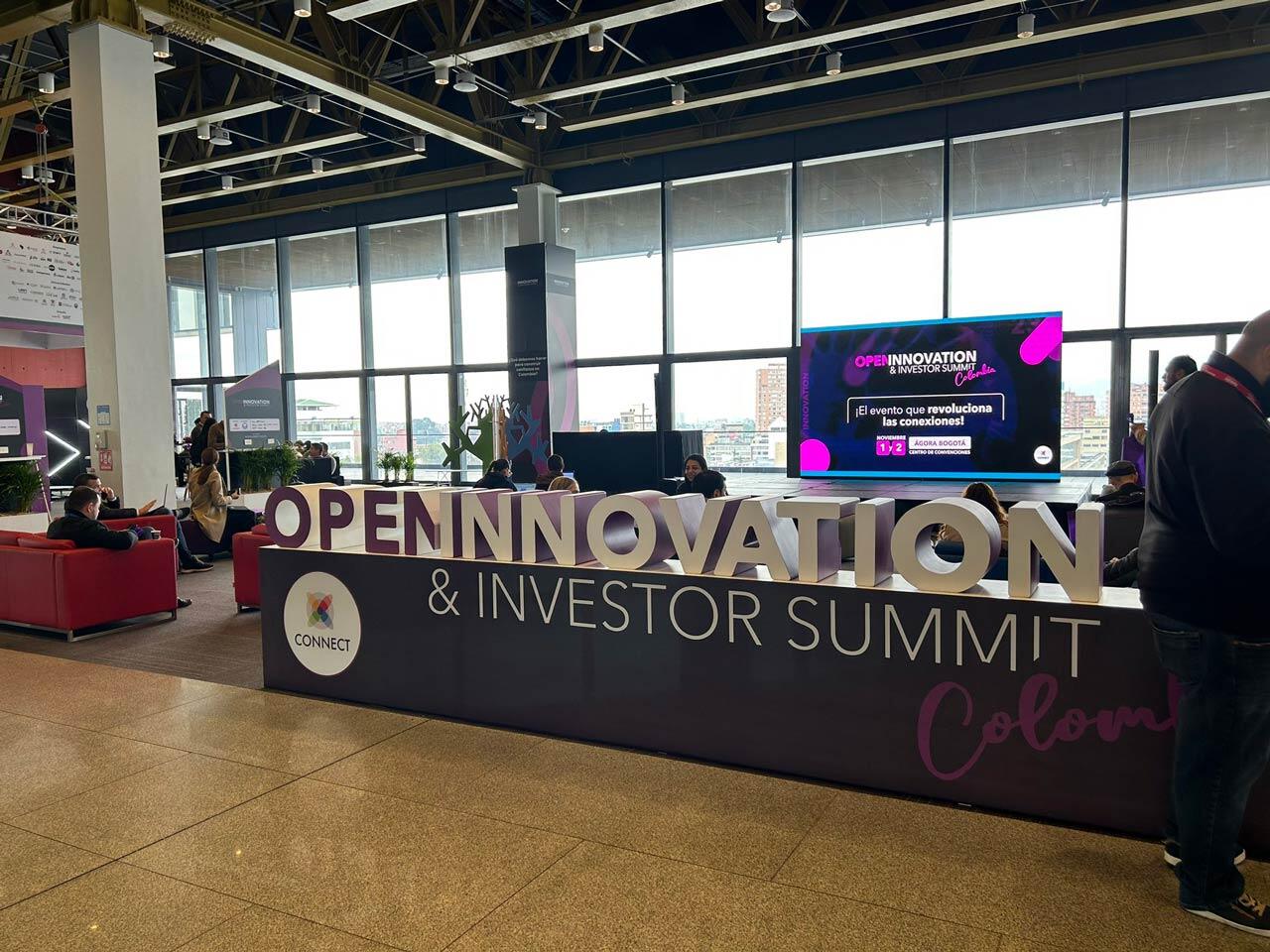 Escenario del evento 'Open Innovation & Investor Summit' con pantallas y asistentes en un espacio moderno con vista a la ciudad.