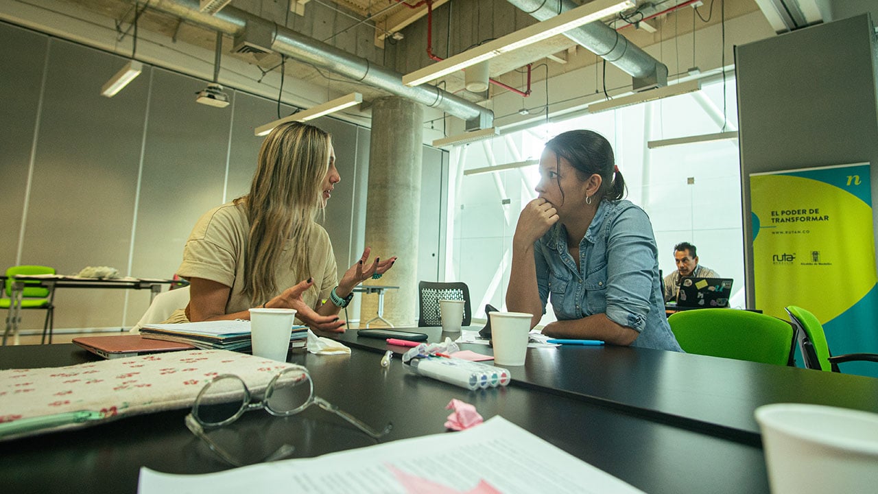 Dos mujeres conversando en una mesa durante una reunión, rodeadas de objetos como tazas y documentos en un espacio moderno y luminoso.