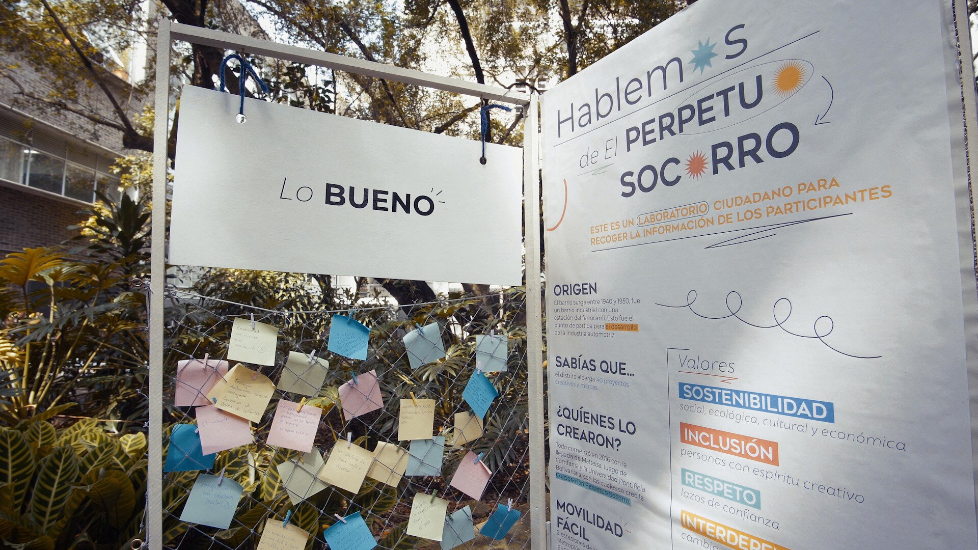 Instalación al aire libre con notas adhesivas bajo un letrero que dice 'Lo Bueno', junto a un cartel sobre 'Hablemos de El Perpetuo Socorro' destacando valores como sostenibilidad e inclusión