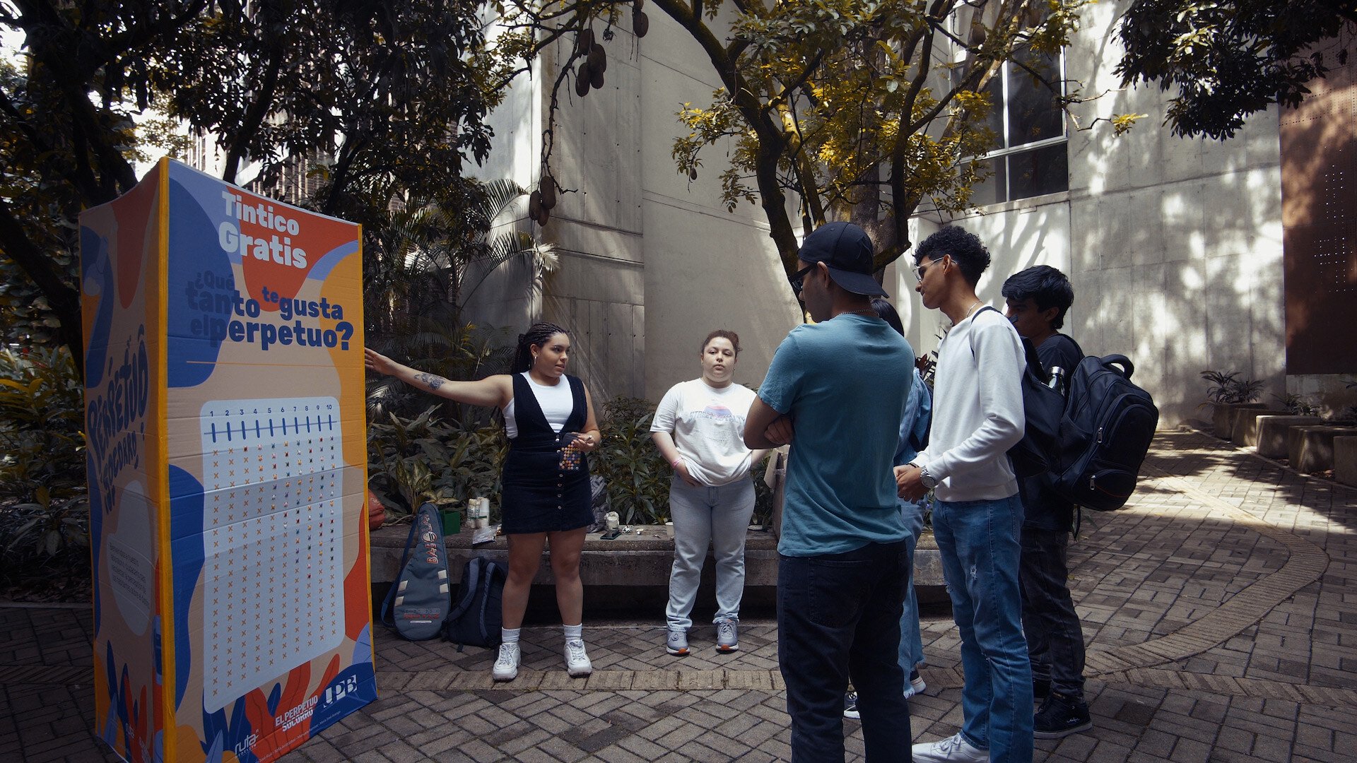 Grupo de personas participando en una dinámica al aire libre junto a un cartel que ofrece 'Tintico Gratis' y plantea la pregunta '¿Cómo te gusta el perpetuo?'