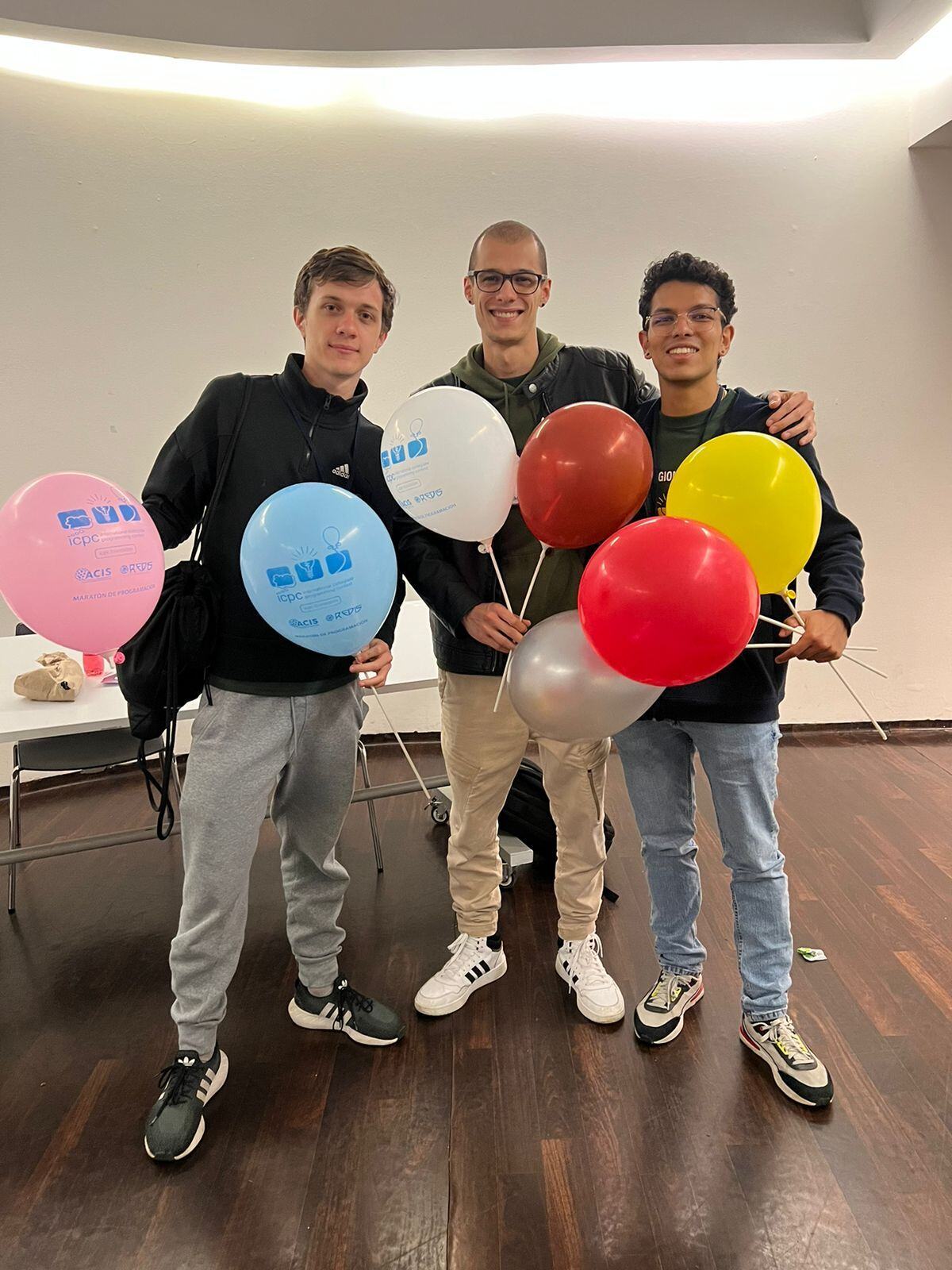 Tres personas sonriendo mientras sostienen globos de colores en un ambiente interior.