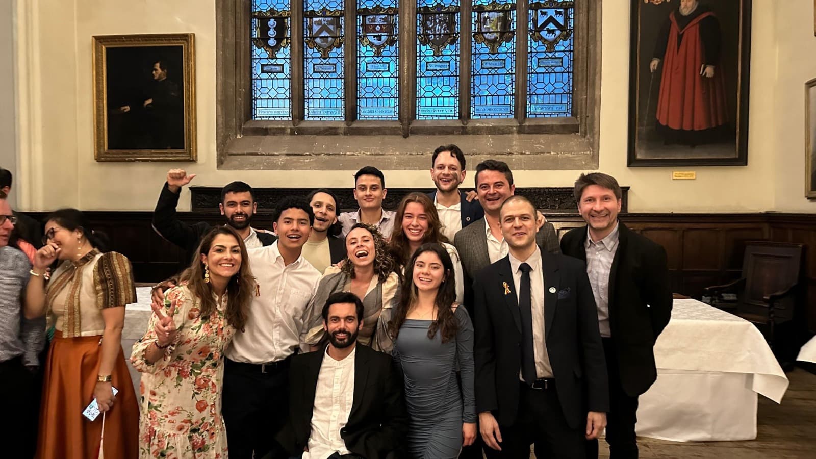 Grupo de personas sonrientes en un evento formal, con un ambiente festivo y una ventana de vitral al fondo.