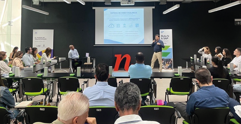 Presentación en un auditorio con una pantalla proyectando información, asistentes sentados y un logotipo rojo en el escenario.