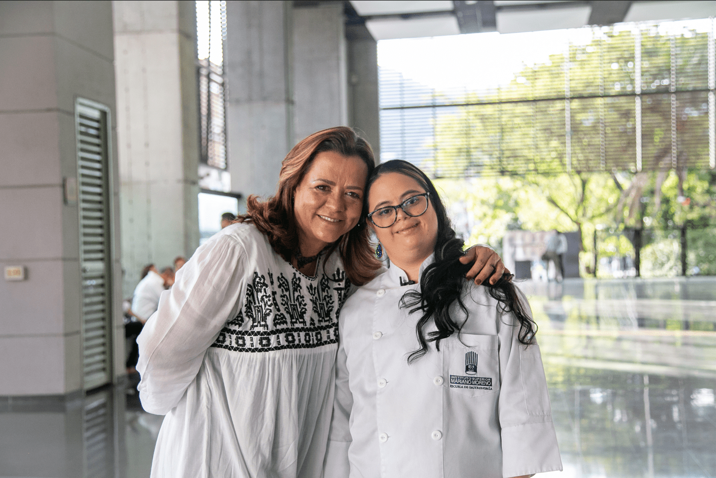 Dos mujeres sonriendo y posando juntas, una de ellas con uniforme de chef, en un espacio moderno y luminoso.