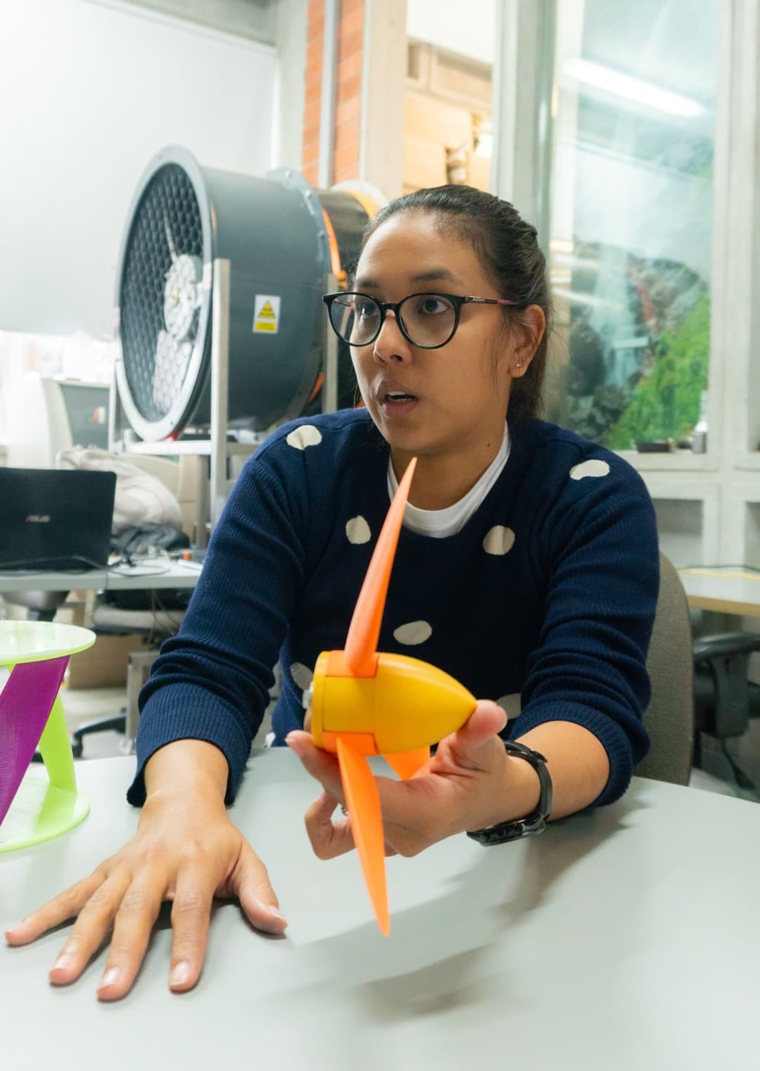 Una mujer explicando el funcionamiento de un modelo de avión hecho de materiales coloridos, mientras lo sostiene en una mesa rodeada de herramientas y otros modelos.