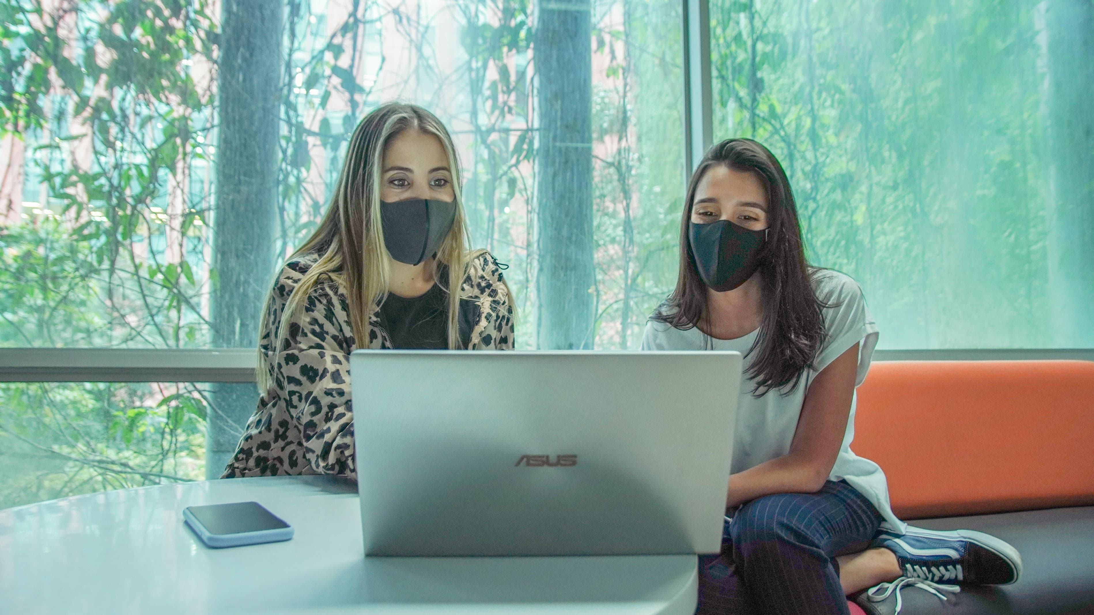 Dos mujeres con mascarillas sentadas frente a un portátil Asus, colaborando en un espacio iluminado con vegetación visible a través de una ventana.