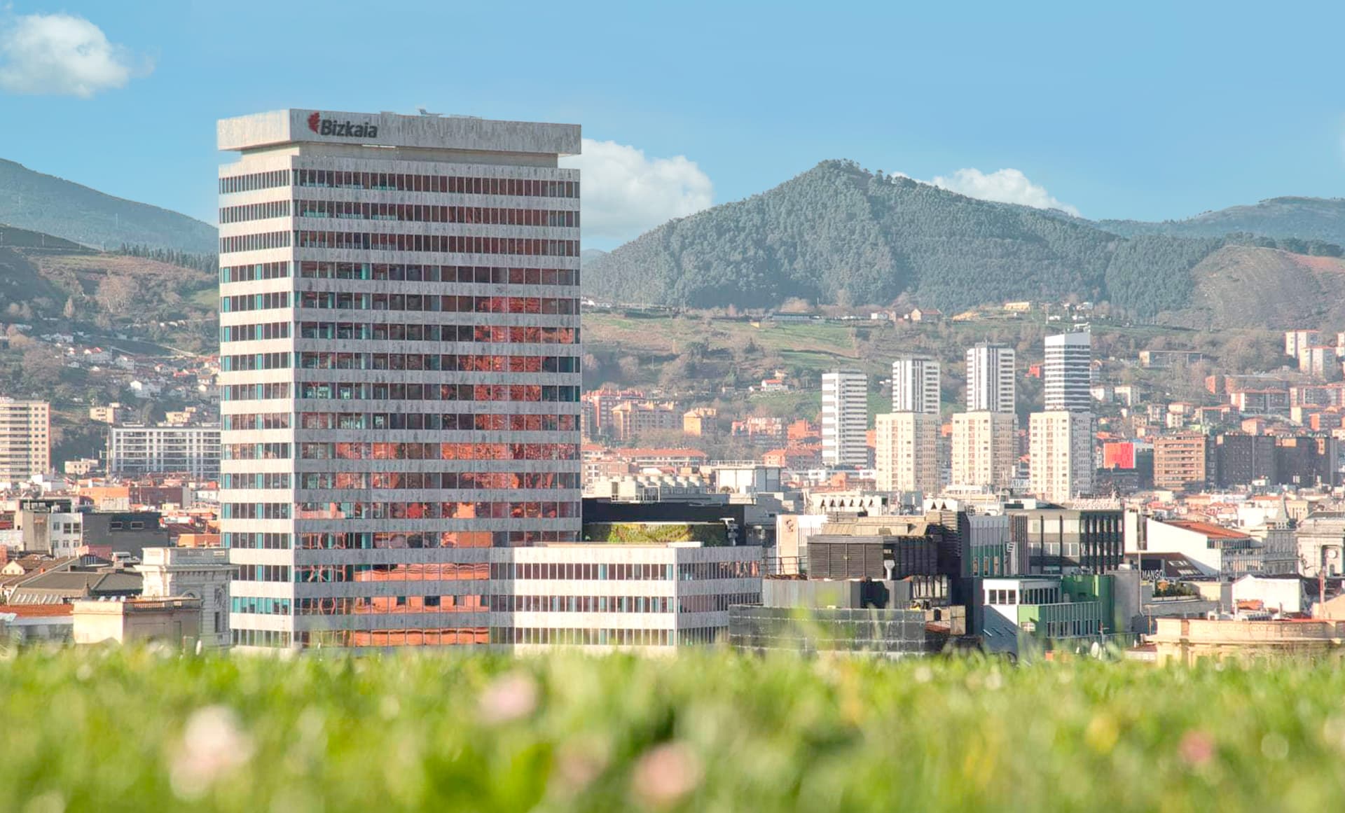 Vista panorámica de una ciudad con un edificio alto destacado en el centro. En el fondo se observan montañas, y la parte frontal muestra césped desenfocado, aportando profundidad a la imagen.