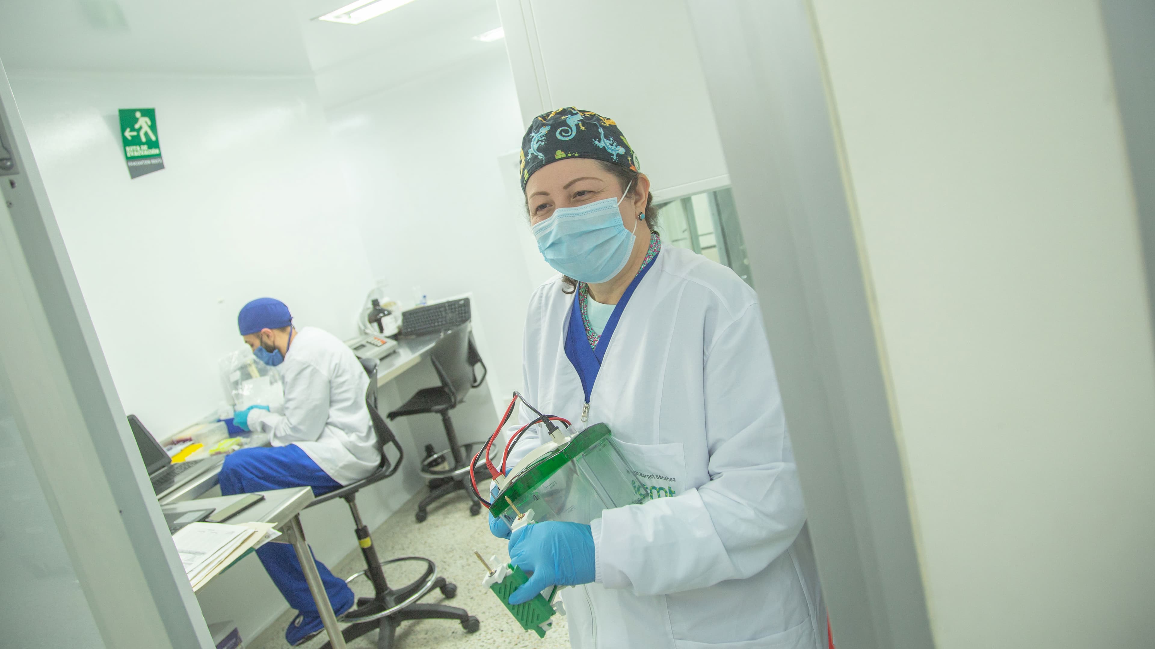 Profesional de la salud con bata, mascarilla y guantes trabajando en un laboratorio, con otro colega al fondo utilizando una computadora.