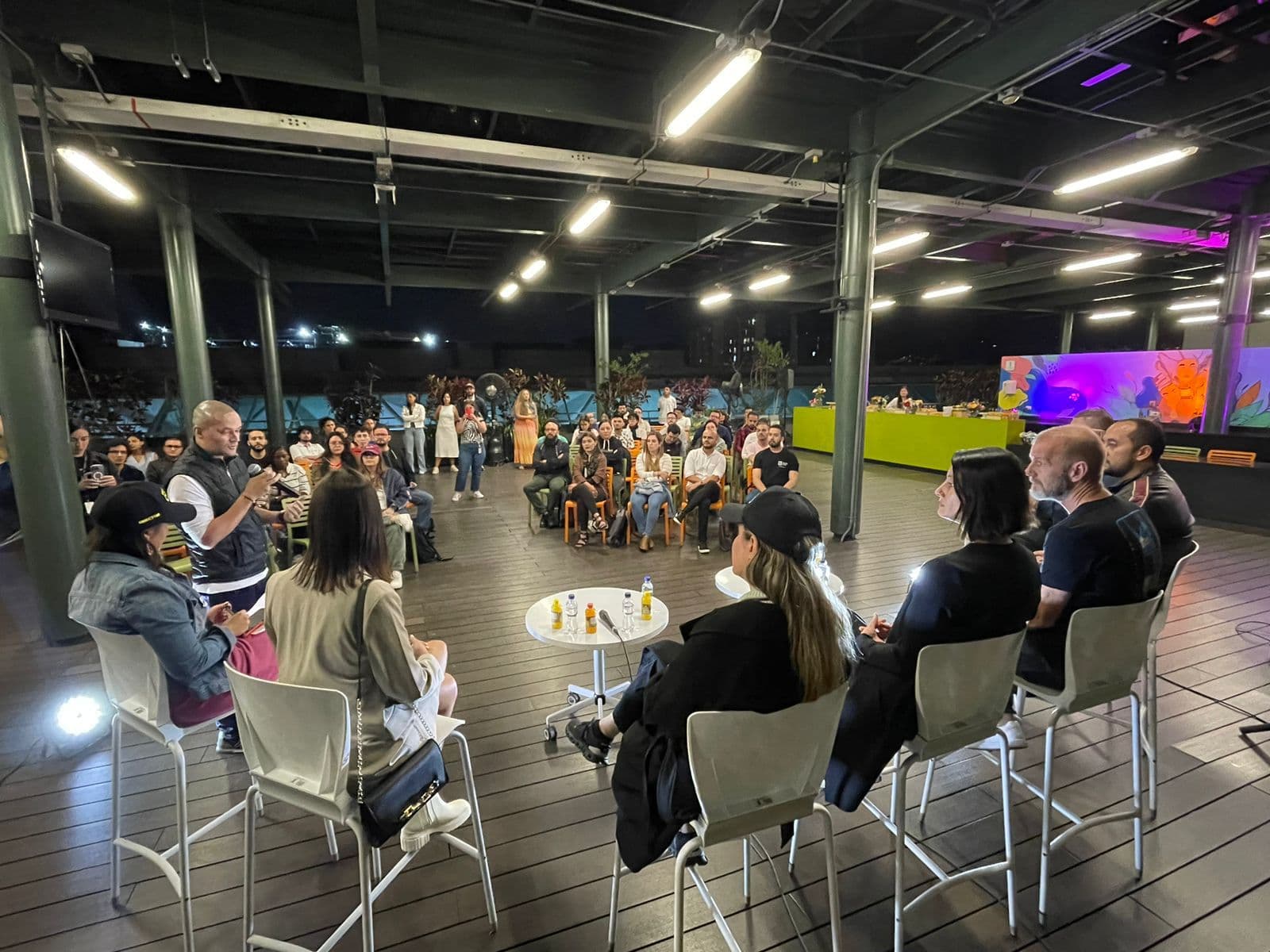 Panel de discusión en un espacio abierto con un público atento y varios panelistas sentados frente a pequeñas mesas circulares