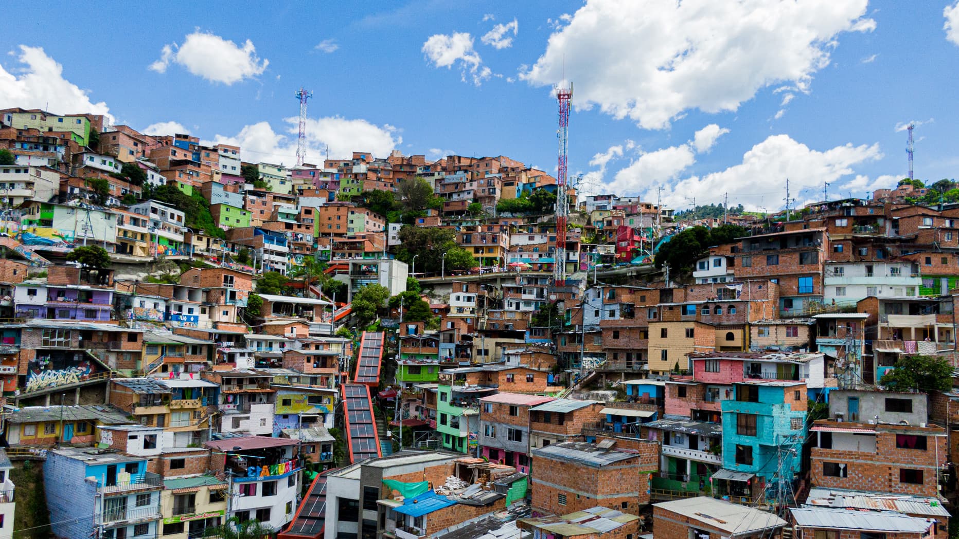 Vista panorámica de un barrio de la Comuna 13 en Medellín con casas coloridas en una ladera, bajo un cielo azul con nubes dispersas.