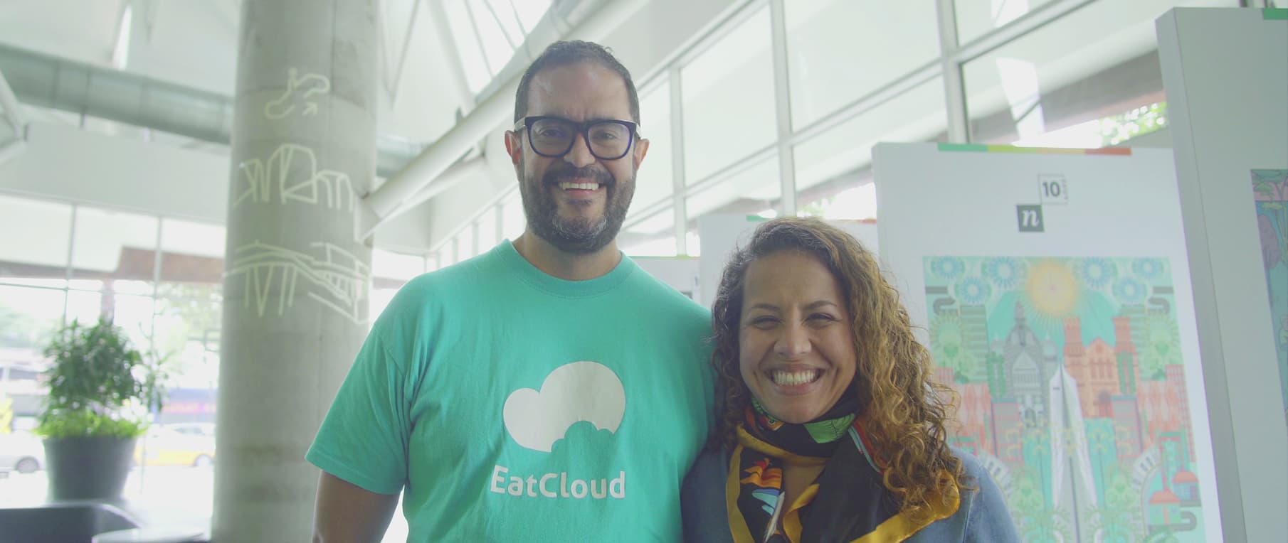 Dos personas sonrientes posando juntas, una de ellas usando una camiseta de EatCloud, en un ambiente luminoso y moderno.