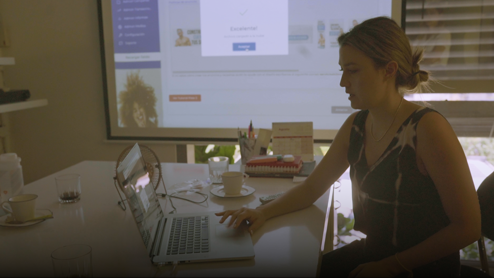 Mujer trabajando en un portátil en una sala de reuniones, con proyección en pantalla al fondo.