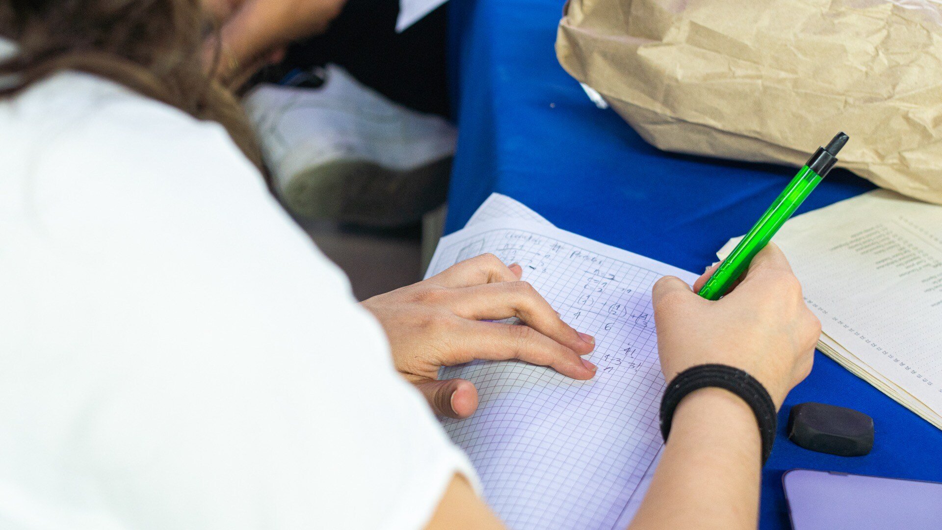 Persona escribiendo en un cuaderno de cuadrícula con un bolígrafo verde, en una mesa azul con papeles y objetos alrededor.