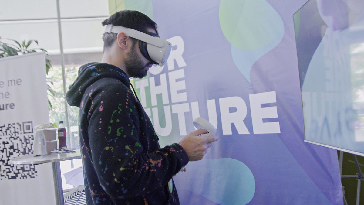 Persona utilizando un visor de realidad virtual con control en mano, frente a una pantalla. Al fondo, se ve un cartel que dice 'FOR THE FUTURE' con un diseño colorido.