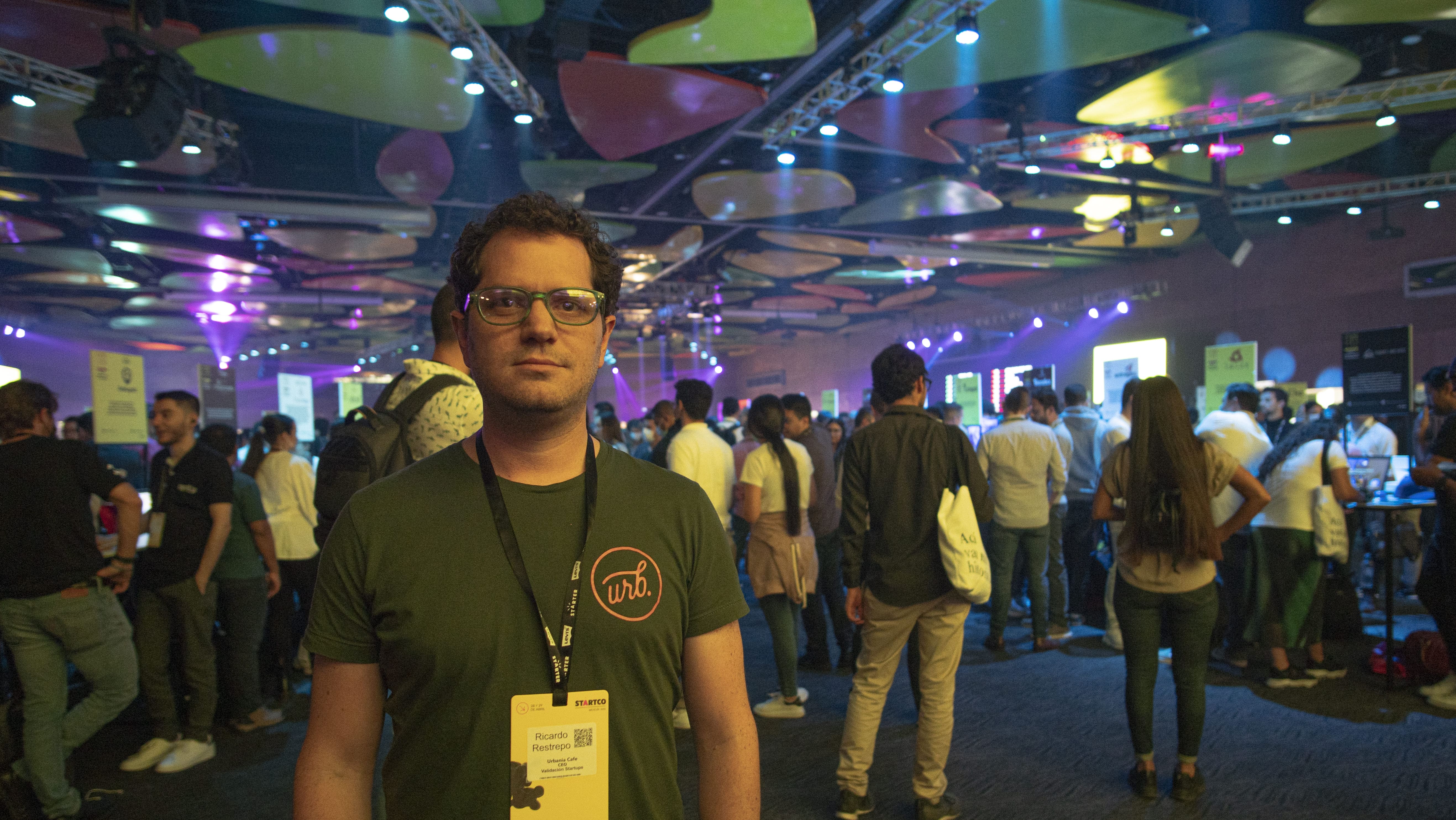 Persona con gafas en primer plano en un evento concurrido con iluminación colorida y un ambiente vibrante.