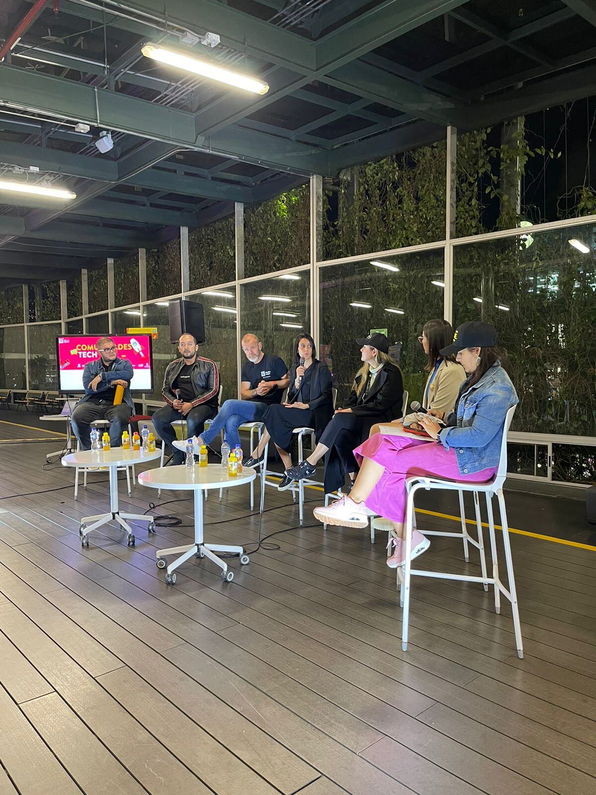 Panelistas sentados en sillas altas participando en una discusión, con mesas pequeñas al frente y una pantalla de fondo en un espacio abierto.