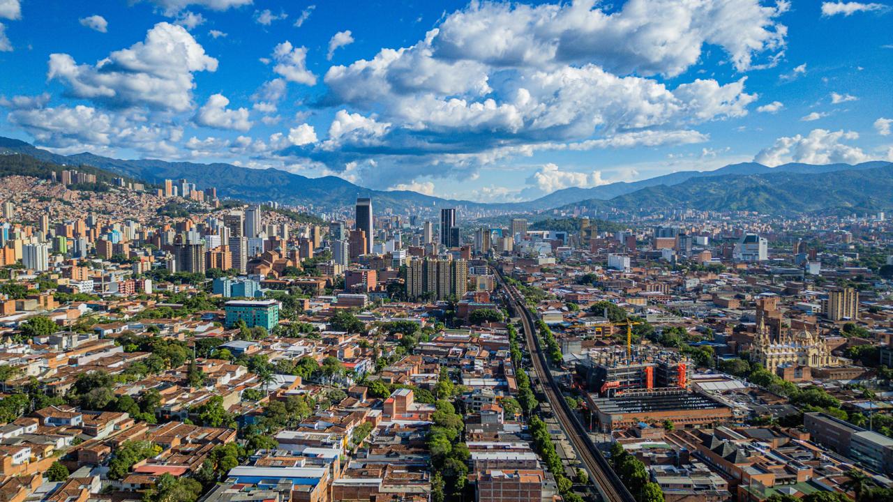 Vista aérea de Medellín con su centro urbano, líneas del metro y montañas al fondo, representando su desarrollo urbano y potencial como ciudad inteligente.